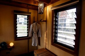 A room with a wooden bench, a robe hanging on a rack, a lamp, and two windows with blinds.