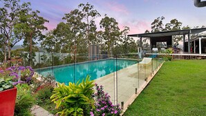 Una piscina con barrera de cristal, rodeada de césped cuidado y diferentes plantas.