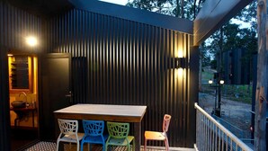 An outdoor patio with a wooden table and colorful chairs, a black wall with vertical slats, and a metal railing. - Amazing Gold Coast Tree House Perfect for Glamping in Queensland (Gold Coast)