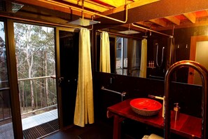 A bathroom with a wooden vanity, a red basin, a shower, and a view of trees through the door.