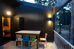 An outdoor patio with a wooden table and colorful chairs, a black wall with vertical slats, and a metal railing.