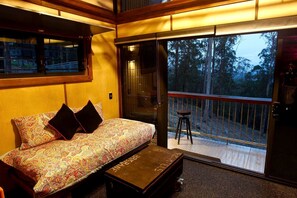 A room with a sofa, a small table, a sliding glass door leading to an outdoor deck, and a window with a view of trees.