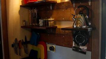 A compact kitchen with a gas stove, shelves with kitchenware, a kettle, and a coffee maker.