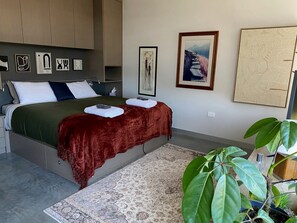 A bedroom with a bed, a bedside table, a large plant, and artwork on the walls.