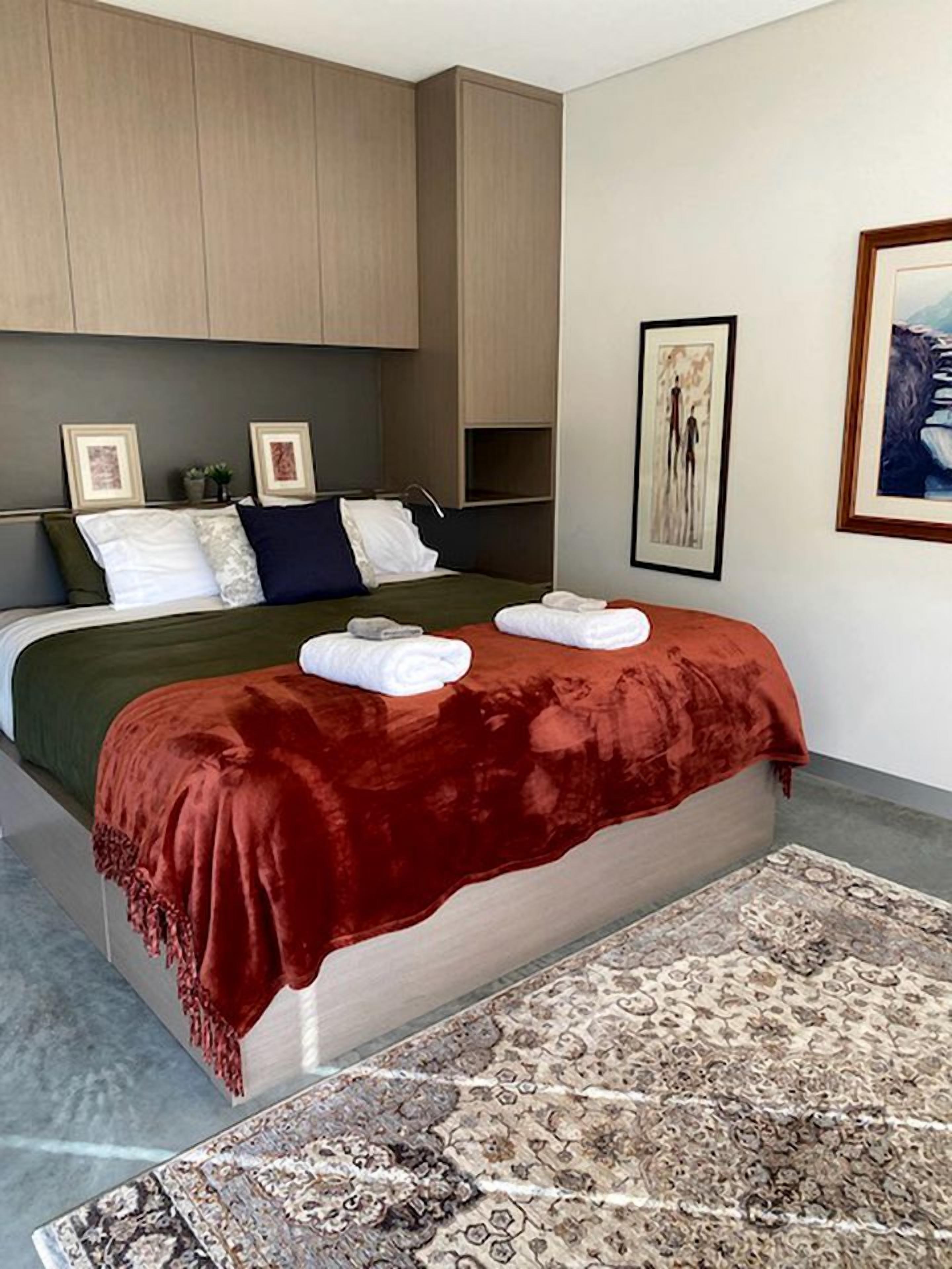 Une chambre à coucher moderne avec un lit, un plaid rouge, une tête de lit et un tableau encadré au mur.