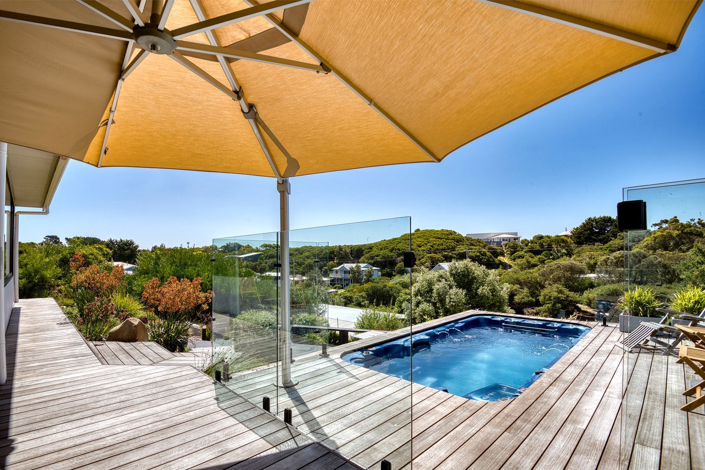 A wooden deck with a glass-enclosed hot tub, a large umbrella, and a view of a hillside with greenery.