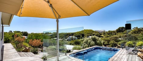 A wooden deck with a glass-enclosed hot tub, a large umbrella, and a view of a hillside with greenery.