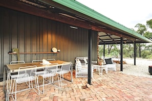 Un patio cubierto con una mesa de madera y sillas de metal blanco, suelo de ladrillo y techo verde.