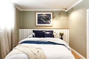 A bedroom with a bed, a framed picture on the wall, a bedside lamp, and a window with curtains.