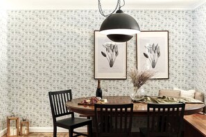 A dining area with a round table, chairs, a pendant light, framed botanical prints, a vase with dried plants, and a bottle of wine.