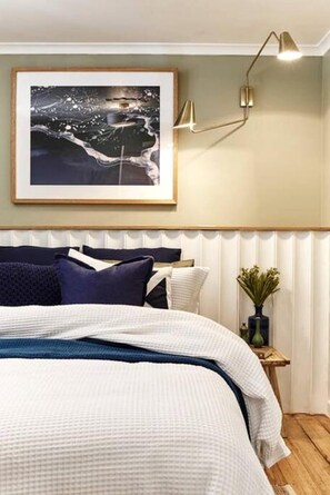 A bedroom with a bed, a framed picture on the wall, a bedside table with a vase, and a wall-mounted lamp.