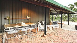 A covered patio area with a wooden table and white metal chairs, a brick floor, and a green roof.
