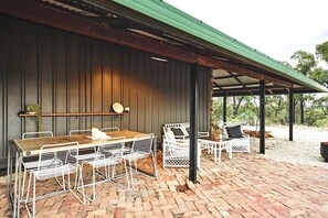 A covered patio area with a wooden table and white metal chairs, a brick floor, and a green roof. - Sweet Off-Grid Cottage with Modern Interior and Exterior in Victoria (Bailieston)