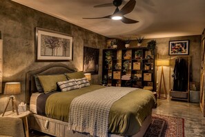 A bedroom with a bed, a ceiling fan, a wall-mounted shelf with plants, a framed picture, and a floor lamp.