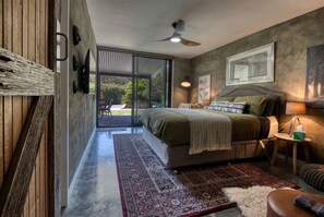A bedroom with a large bed, a view of the outdoors, a ceiling fan, and a nightstand with a lamp.