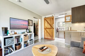 A modern living room with a flat-screen TV, bookshelves, a coffee table, and a kitchen area with cabinets and a countertop.