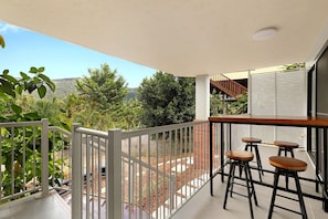 Ein Balkon mit weißer Reling, einem Holztisch und drei Barhocker.