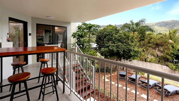 A balcony with a wooden table and stools, a railing, and a view of a garden with trees and a hilly landscape.