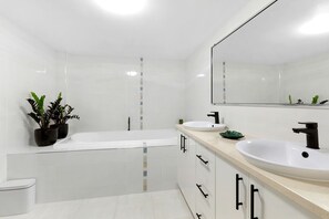 A modern bathroom with a bathtub, two sinks, a toilet, and potted plants.