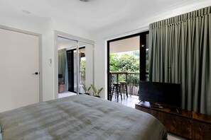A bedroom with a bed, a TV, a balcony with a table and chairs, and a sliding door leading to an outdoor area.