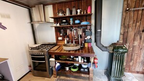 A compact kitchen with a gas stove, oven, and wooden shelves holding various kitchen utensils and cookware.