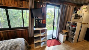 A room with a bed, a TV on a stand, a small table, a refrigerator, and a window with a view of trees.