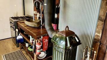 Una cocina rústica con una estufa de leña, repisas con utensilios de cocina y un tapete estampado.