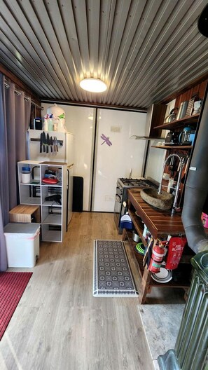 A compact kitchen with a built-in stove, a small refrigerator, and a wooden shelf with kitchen utensils.