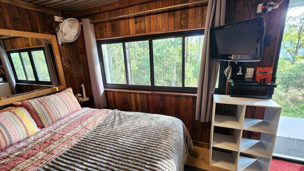 A bedroom with a bed, a TV, a shelf, and a window with a forest view.