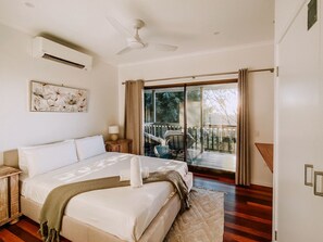 A bedroom with a bed, bedside tables, a ceiling fan, an air conditioning unit, a large window with curtains, and a view of an outdoor deck. - Secluded Mountain Cabin with Private Sauna & Ice-Bath Near Springbrook, Mudgeeraba, Queensland (Mudgeeraba)