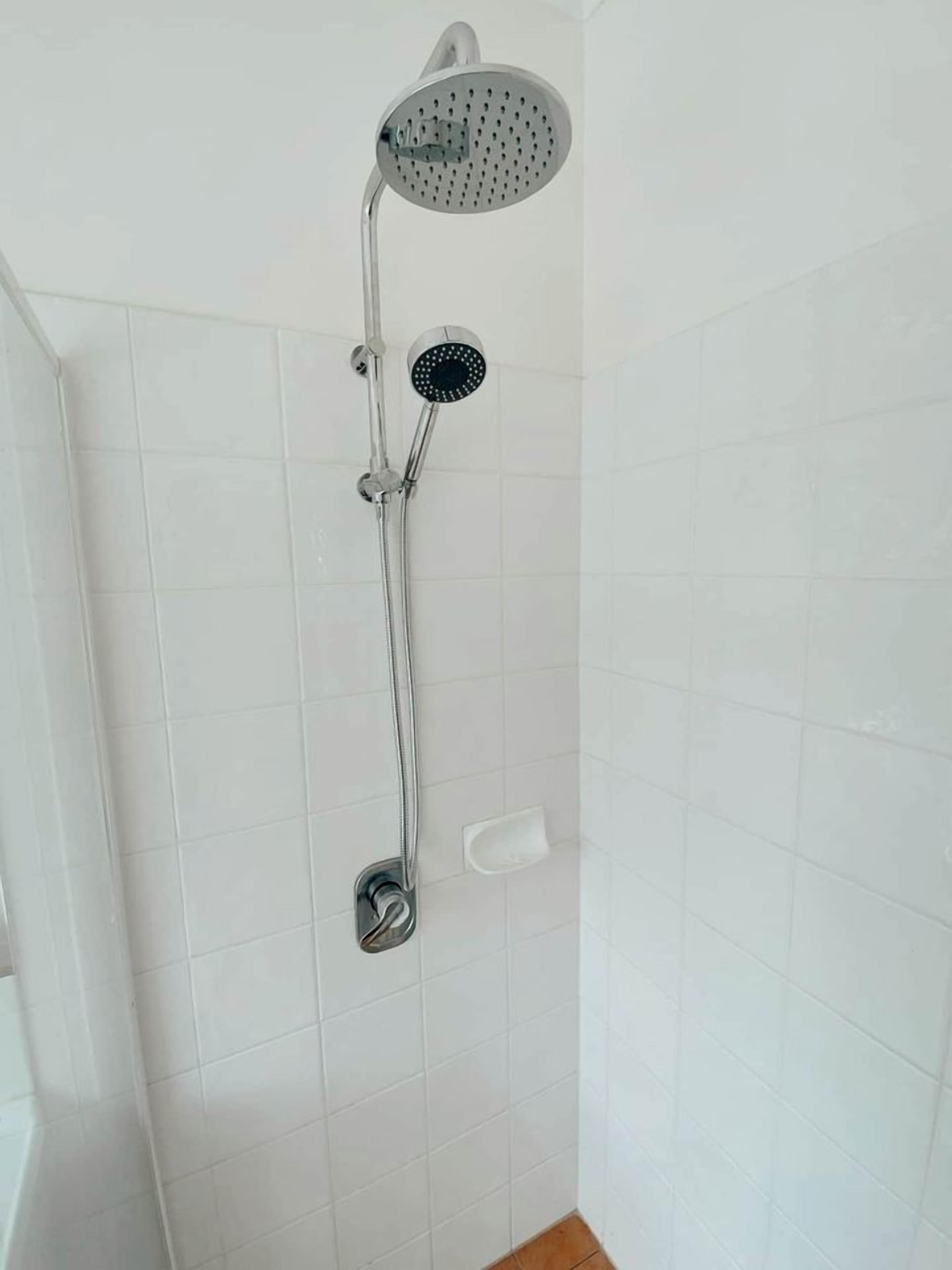 Une douche équipée d’un pommeau de douche à main et d’un pommeau de douche mural, dans une salle de bain aux murs recouverts de carreaux blancs.