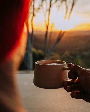 Eine Person hält eine Tasse, aus der Dampf aufsteigt, was auf ein warmes Getränk hindeutet. Im unscharfen Hintergrund sind Bäume und ein Sonnenuntergang zu sehen.