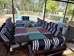 Un patio exterior con piscina, rodeado por una valla blanca y amueblado con cojines a rayas y una mesa.