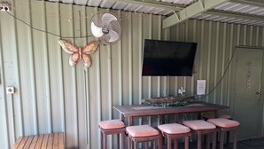 Un área de estar al aire libre con techo, que incluye una mesa, taburetes de bar, un ventilador y un televisor en la pared.
