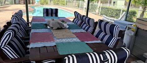 Une terrasse extérieure avec une piscine, entourée d’une clôture blanche et aménagée avec des coussins à rayures et une table.