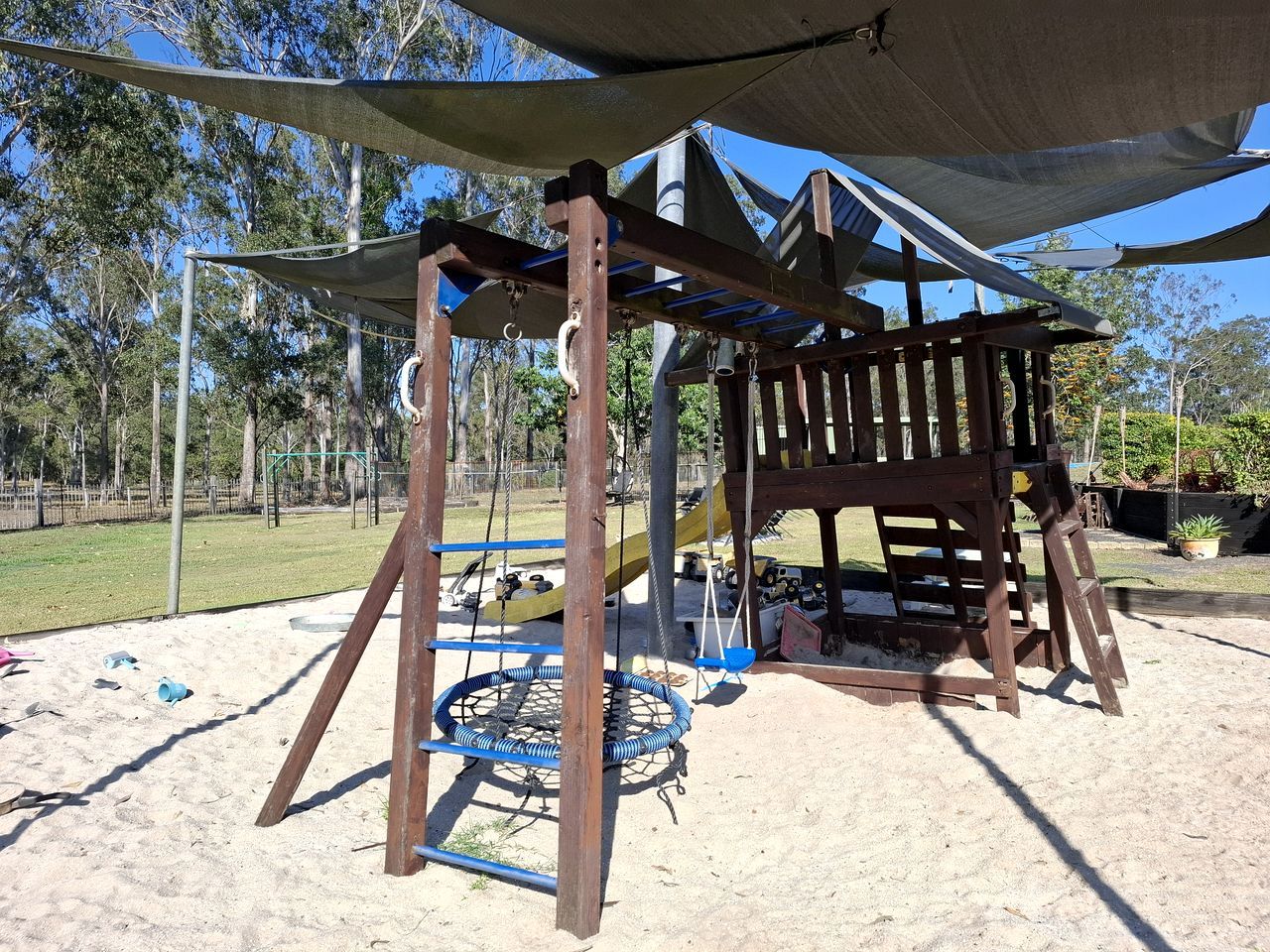 A wooden play structure with a swing set and a shaded area, surrounded by sand and trees.