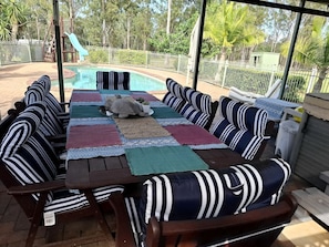 An outdoor patio with a pool, surrounded by a white fence, and furnished with striped cushions and a table.