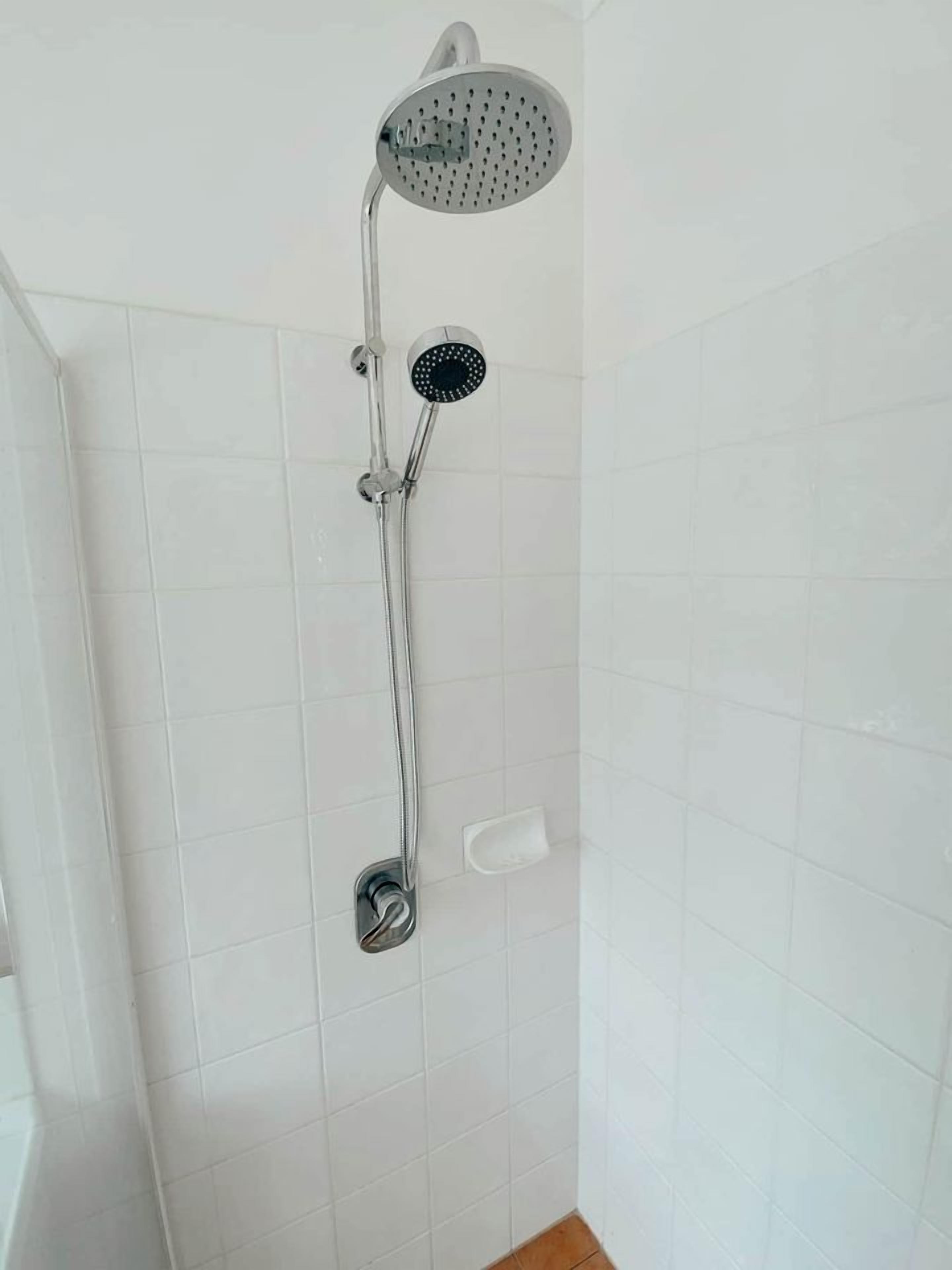Une douche équipée d’un pommeau de douche à main et d’un pommeau de douche mural, dans une salle de bain aux murs recouverts de carreaux blancs.