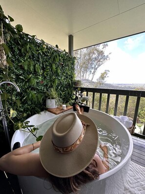 Eine Person entspannt in einer Hot Tub, mit Hut, umgeben von üppiger Vegetation und Blick ins Freie.