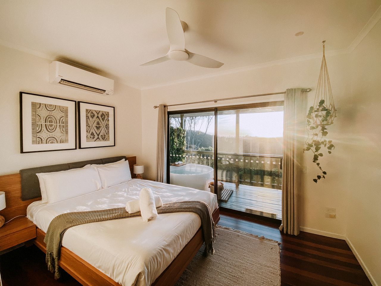 A bedroom with a large bed, a ceiling fan, an air conditioning unit, and a balcony with a view.