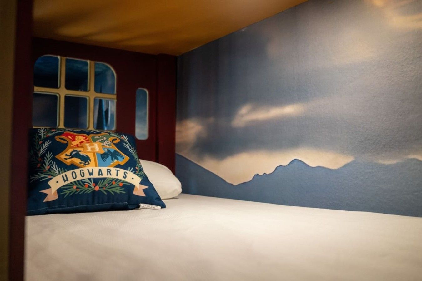 A bedroom with a bed, a pillow with a Hogwarts logo, a window, and a mountain landscape mural on the wall.