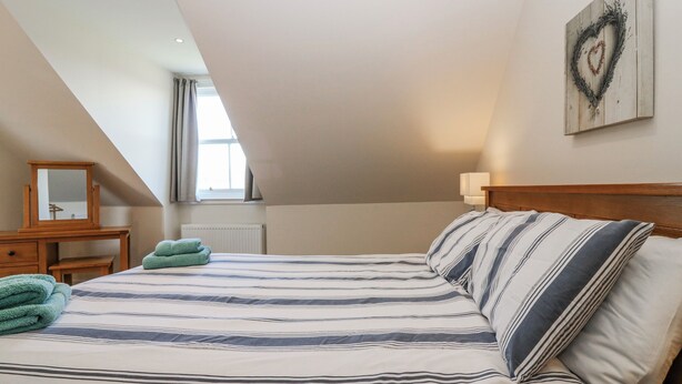 Ein Schlafzimmer mit schrägem Dach, einem Bett mit gestreifter Bettwäsche, einer hölzernen Kommode, einem Spiegel und einer herzförmigen Dekoration an der Wand.