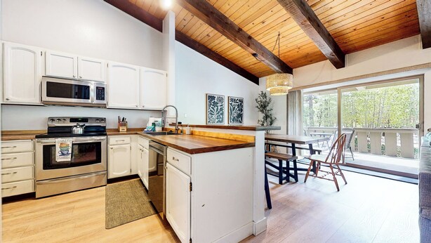 Eine Küche mit weißen Schränken, einer Mikrowelle, einem Backofen und einem Spülbecken. Es gibt eine Holzbalkendecke, einen Essbereich mit Tisch und Stühlen sowie einen Blick durch große Fenster.