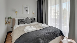 Un lit bien fait, recouvert d’une courtepointe, une table de chevet avec une lampe, un tableau encadré au mur et une fenêtre avec des rideaux.