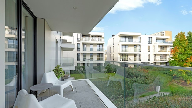 Un balcon avec des chaises blanches et une petite table, donnant sur un jardin et les bâtiments adjacents.