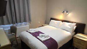 Habitación de hotel con cama, una silla, mesita de noche, lámpara y ventana con cortinas.