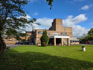 A building with a brick facade, a flat roof, and a small tower. There is a paved area in front with a tree and a parked motorcycle.
