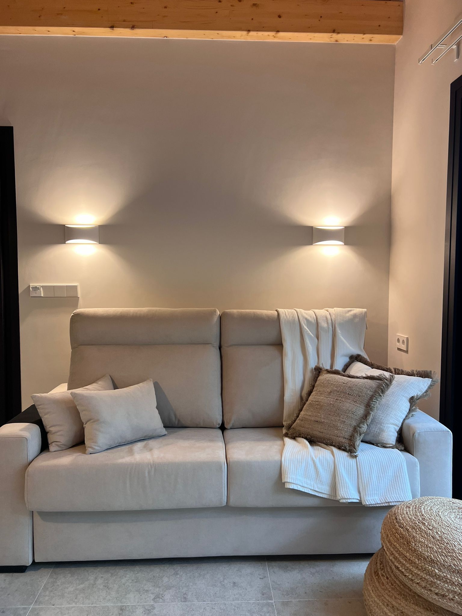 Een moderne woonkamer met een beige bank, twee wandlampen en een geweven poef.