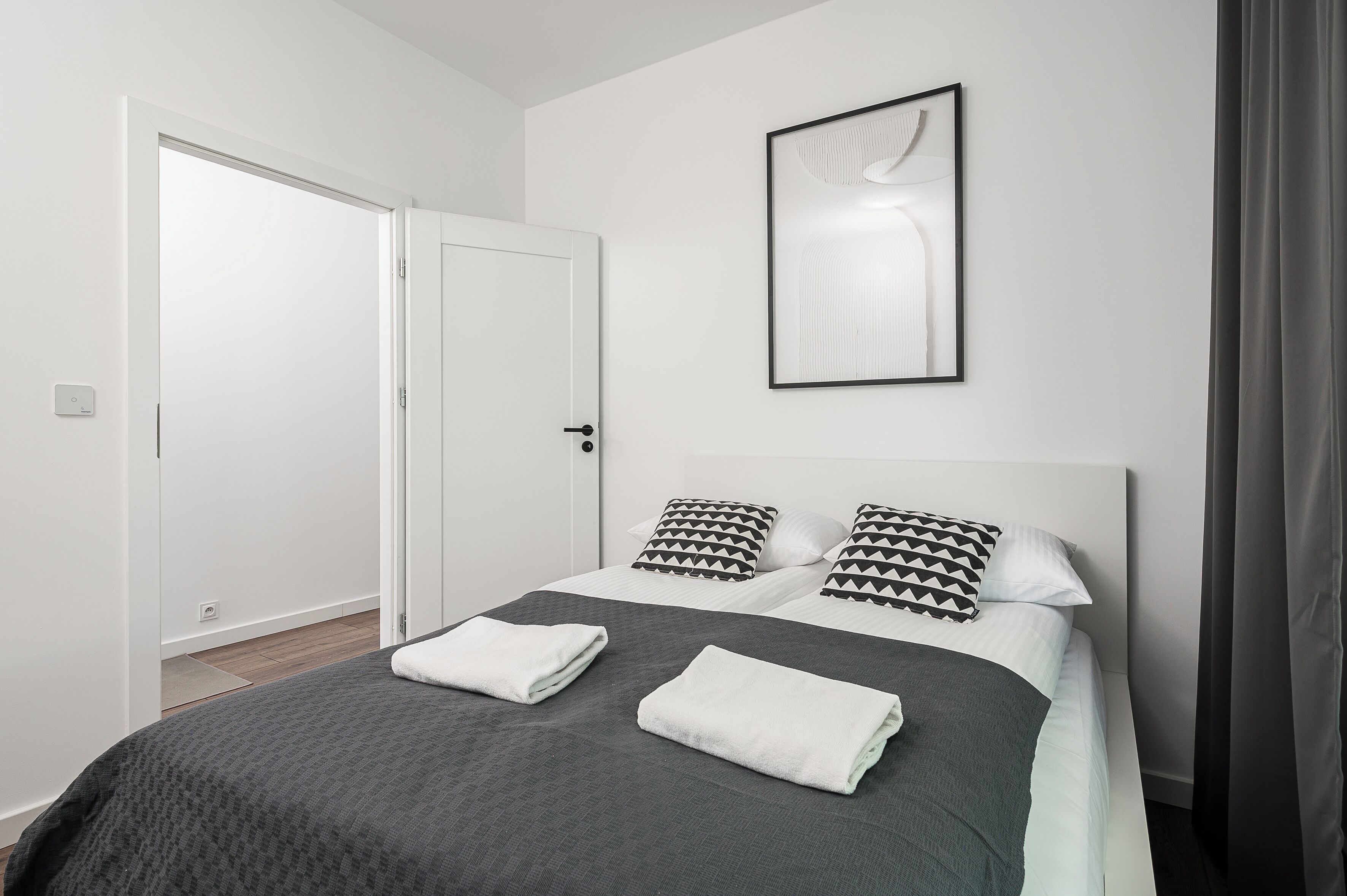 A modern hotel room with a neatly made bed, a framed artwork on the wall, and a door leading to another room.