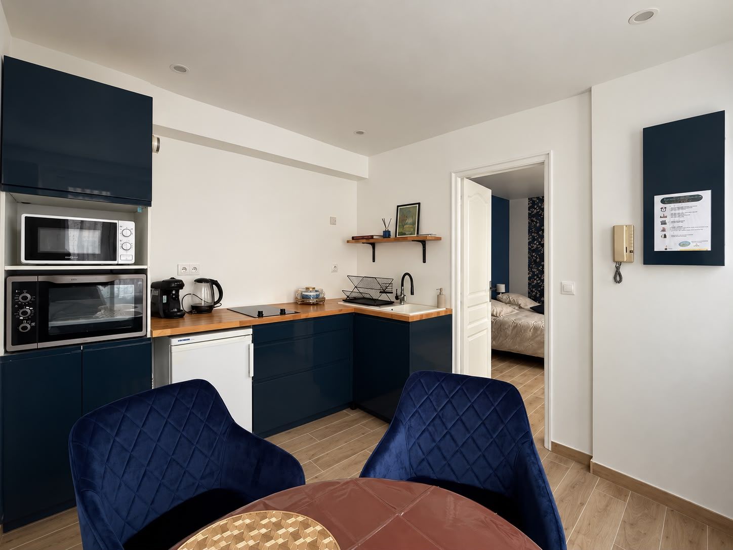 A compact kitchen with modern appliances, a dining area with a table and chairs, and a visible bedroom with a bed.